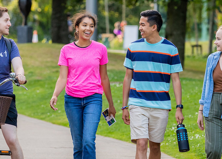 Five students walk together in a grassy campus. One has a bicycle.